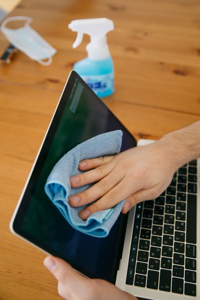 about-01 Une personne nettoie l’écran d’un ordinateur portable avec un chiffon bleu et un vaporisateur sur une table en bois pour des raisons d’hygiène.